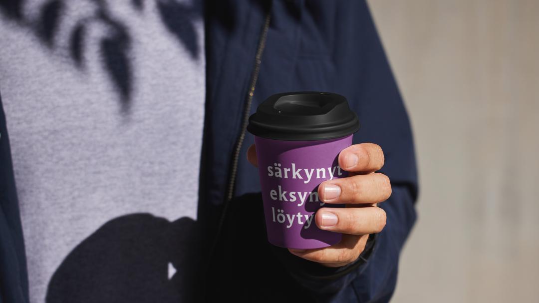 Mies pitää violetista kahvikupista, jossa on teksti 